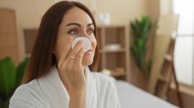 Genç, esmer saçlı bir kadın spa odasında bornoz giyerken cilt bakımı uyguluyor. Etrafı iç güzellik ve güzellik unsurlarıyla çevrili..