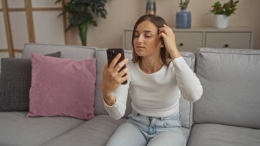 Genç, güzel, sarışın bir kadın oturma odasında oturuyor, akıllı bir telefon kullanıyor, rahat bir kanepeye yaslanıyor, modern, rahat bir ev içinde..