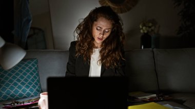 Konforlu bir oturma odasında dizüstü bilgisayarıyla geç saatlere kadar çalışan odaklanmış genç bir kadın, modern telekomünikasyon....
