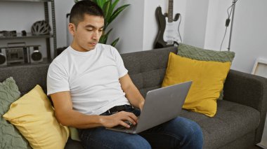 Evdeki gri koltukta otururken, arka planda gitar çalan günlük giysili İspanyol bir adam dizüstü bilgisayarda çalışıyor..