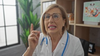 Olgun sarışın kadın doktor, gözlük ve beyaz önlük takıp arka planda bitki ve raflarla modern bir klinikte dururken parmağıyla işaret ediyor..