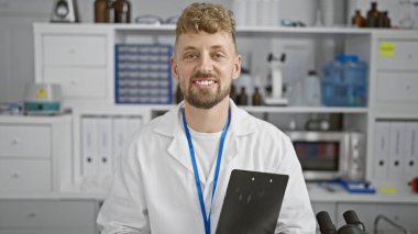 Mavi gözlü, sakallı, laboratuvar önlüğü ve ipiyle laboratuvar ortamında elinde bir pano tutan yakışıklı bir genç..