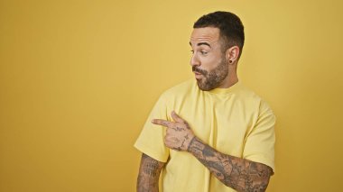 Sakallı ve dövmeli yakışıklı İspanyol bir adam sarı izole bir arka planı işaret ediyor.