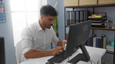 Modern bir ofiste çalışan yakışıklı genç adam arka planda klasörleri ve rafları olan bir masada otururken bilgisayarda yazı yazıyor.