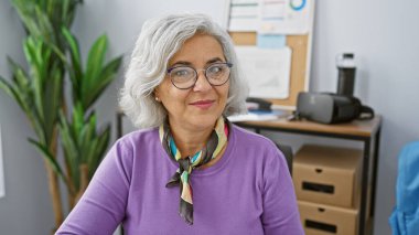 Ofis ortamında üzerinde bitkiler ve teknolojik aletlerle gözlük takan gülümseyen gri saçlı kadın..