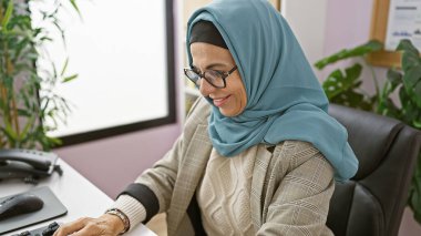 Profesyonel, orta yaşlı, gözlüklü ve tesettürlü bir kadın bilgisayarında çalışıyor..