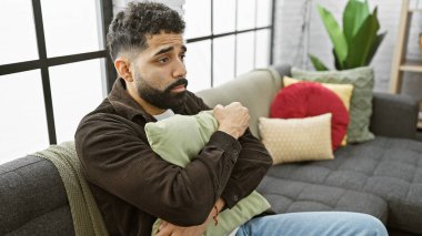 Modern bir oturma odasında oturan sakallı, düşünceli İspanyol adam yalnızlığı yansıtıyor..