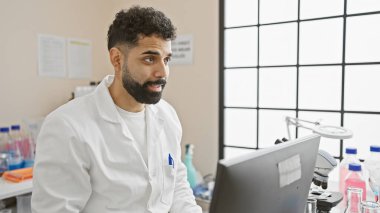 Modern laboratuvarda bilgisayarla çalışan, labrador giymiş yakışıklı İspanyol bir adam.