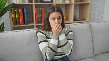 İspanyol kadın koltukta şoke olmuş bir şekilde oturuyor. Kapalı yerde ağzı ellerle kapatıyor.