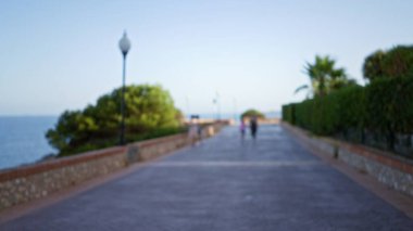 Defocused image of a blurred outdoor pathway with people walking, surrounded by greenery, ocean view, lampposts, and clear sky. clipart