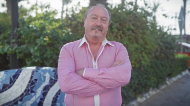 Çapraz kollu, kendinden emin orta yaşlı bir adam yeşil ve kentsel geçmişi olan bir parkta gülümsüyor..