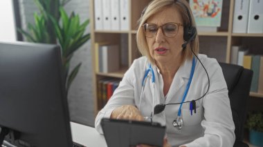 Bir klinikte yaşlı bir kadın doktor, kulaklık takıyor ve beyaz önlük giyiyor, kapalı alanda bir tablet bilgisayar üzerinde çalışıyor..