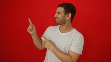 Beyaz tişörtlü çekici genç adam izole edilmiş kırmızı arka plan üzerinde zıt yönleri işaret ediyor.