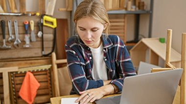 Aletler ve ahşap mobilyalarla çevrili bir ahşap atölyede dizüstü bilgisayar kullanan odaklanmış sarışın kadın..