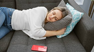 Beyaz tenli sarışın genç bir kadın, evinde rahat bir kanepede, yanında yastıklar ve akıllı bir telefonla uyuyor..