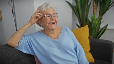 Happy senior woman with grey hair and glasses laughing while relaxing on a sofa at home clipart