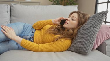 Genç ve çekici sarışın kadın oturma odasında kanepede uzanıyor, telefonda konuşuyor, günlük kıyafetler giyiyor..