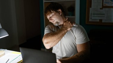 Genç, sakallı, sarışın bir adam boynunu tutarken acı içinde kıvranıyor ve loş bir ofis odasında dizüstü bilgisayarın başında çalışıyor..