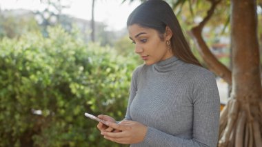 Gri balıkçı yakalı genç bir İspanyol kadın telefonunu güneşli, yemyeşil bir bahçede kullanıyor..