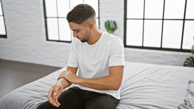 Yakışıklı sakallı adam minimalist bir yatak odasında düşünceli bir şekilde yatakta oturuyor..