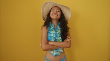 Mavi bikinili ve güneş şapkalı Asyalı bir kadın sarı arka planda çapraz kollarla gülümsüyor, kendine güveni ve yaz titreşimleri yayıyor..