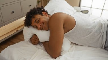 Genç, ispanyol, yatak odasında yatakta yatan bir adam, evde rahat ve mutlu görünüyor.