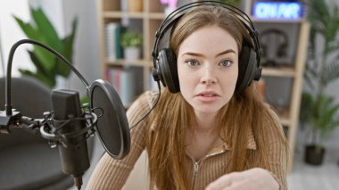 Kulaklıklı genç bir kadın modern bir radyo stüdyosunda mikrofona konuşuyor..