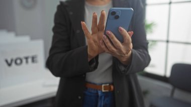 Olgun bir kadın, kapalı bir seçmen odasında oy verme kabinleriyle çevrili dururken akıllı telefonunu kontrol eder..