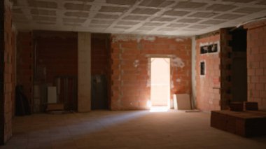 Tamamlanmamış bir inşaat sahasının içi bulanık tuğla duvarlar, dağınık yapı malzemeleri ve bir kapı aralığından geçen doğal ışık.