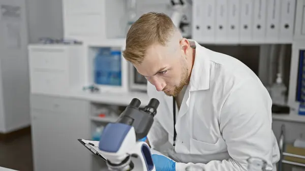 stock image A young bearded man in a laboratory observes a microscope while holding a clipboard.