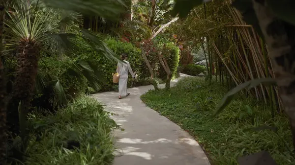 Genç bir kadın, tropikal bir ortamda huzur ve rahatlığı somutlaştıran yemyeşil bir Bali tatil köyünde yürüyor..