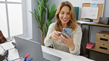 Düzenli ofis masasında akıllı telefon kullanan gülümseyen genç bir kadın pozitifliğini ve memnuniyetini ifade ediyor..