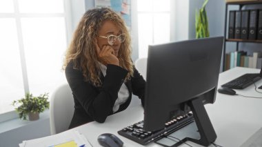 Ofiste çalışan İspanyol kadın, bilgisayar ekranına odaklanmış, organize belgeler ve dosyalarla profesyonel ve modern bir iş ortamı yansıtıyor.