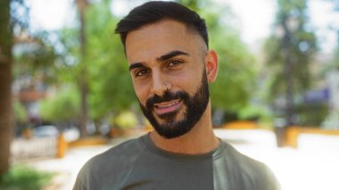 Güneşli bir günde şehir parkında gülümseyen sakallı yakışıklı genç bir adam. Arka planda yeşil ağaçlar bulanık.