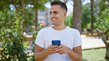 Neşeli, genç, İspanyol bir adam zengin bir şehir parkında güneşli havanın tadını çıkarırken akıllı telefonuyla mesaj atıyor..