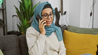 Orta yaşlı, tesettürlü ve gözlüklü bir kadın rahat bir oturma odasında akıllı bir telefonla konuşuyor..