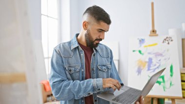 Kot ceketli genç sakallı bir adam arka planda resimler olan parlak bir sanat stüdyosunda dizüstü bilgisayar kullanıyor..