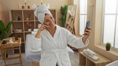 Kaplıcada selfie çeken bir kadın, beyaz bornoz ve havlu giyiyor, rahat ve güzel görünüyor, huzur verici bir sağlık merkezinde, kapalı bitkilerle ve rahatlatıcı bir ambiyansla.