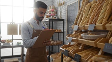 Bir pastanede çalışan sakallı genç İspanyol bir adam. Kapalı bir ortamda çeşitli ekmek somunlarıyla çevrili bir panoya notlar yazıyor.