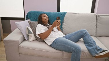 Afrikalı Amerikalı bir kadın modern bir oturma odasında akıllı telefonuyla koltukta dinleniyor..