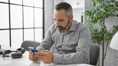 Gri saçlı, olgun sakallı bir adam akıllı telefonunu modern ofis ortamında inceliyor..