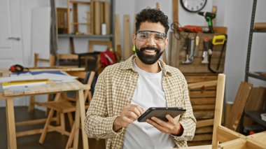 Güvenlik gözlüğü takan sakallı bir adam, iyi donanımlı bir ahşap atölyesinde elinde tablet tutarken gülümsüyor..