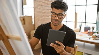 Handsome hispanic man using tablet in art studio with easel and paints clipart