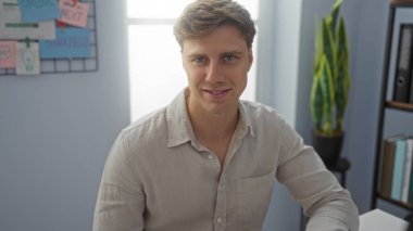 Ofis ortamında rahat bir ifadeyle oturan genç adam profesyonel ama sıradan bir görünüm sergiliyor..