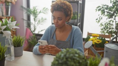Çiçekçide yeşil bitkiler ve çiçek aranjmanları ile çevrili akıllı telefon kullanan Afrikalı Amerikalı bir kadın..