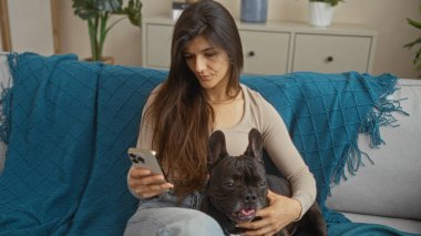 Genç bir İspanyol kadın oturma odasında Fransız bulldoguyla rahat bir kanepede rahatlarken akıllı telefonunu kontrol ediyor..