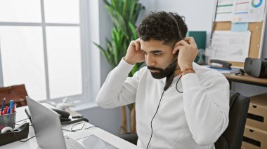 Modern bir ofiste kulaklık takan düşünceli İspanyol bir adam dikkatle bilgisayar ekranına odaklanıyor..