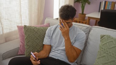 Oturma odasında telefon tutarken gergin görünen genç bir adam koltukta oturuyor.