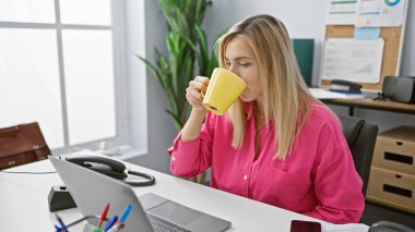 Pembe gömlekli sarışın kadın ofis masasında laptoplu ve telefonlu kahve içiyor.