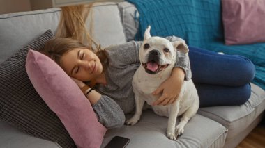 Sarışın, beyaz bir kadın kanepede rahat rahat oturur, rahat bir oturma odasında köpeğine sarılır..